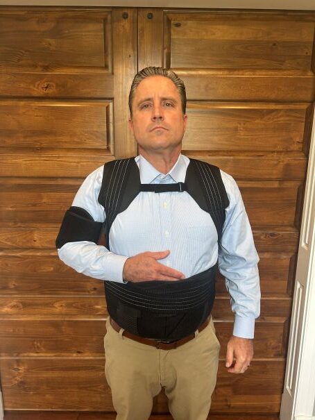 Man wearing a black health support vest with adjustable straps, standing against a wooden background, demonstrating proper fit and functionality.